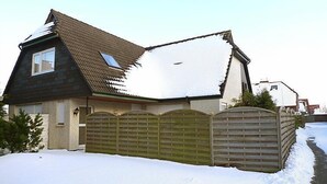 Una casa con techo inclinado, cerca de madera y nieve en el suelo.