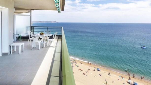 Un balcone con sedie e tavolo bianchi, affacciato su una spiaggia con persone e una barca in acqua.