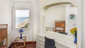 Una camera da letto con un'ampia finestra vista spiaggia, un comodino con una lampada, una cassettiera e uno specchio che riflette l'ambiente.