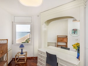 Een slaapkamer met een groot raam dat uitkijkt op een strand, een nachtkastje met een lamp, een dressoir en een spiegel die de kamer weerspiegelt.