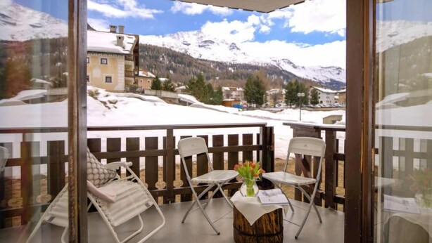 A balcony with a view of snow-covered mountains, two white chairs, and a small wooden table with a vase of flowers. - Chesa Linaria by Interhome (Silvaplana-Surlej)