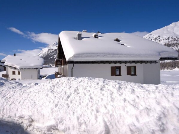 A snow-covered chalet with a balcony, surrounded by mountains and a clear sky.
