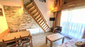 A cozy living room with a wooden staircase, a dining table with chairs, a sofa with patterned cushions, and a TV mounted on the wall. - Clos du Savoy by Interhome (Chamonix)