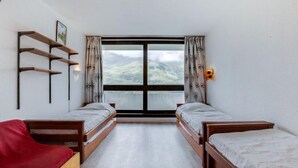 Et værelse med to træsenge, en rød stol, en træhylde med et billede og et vindue med gardiner.
