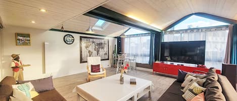 Un salon spacieux avec un plafond haut, des poutres apparentes, une grande télévision et un coin repas comprenant une table et des chaises.
