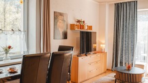 A modern living room with a dining area, a large window with curtains, a painting on the wall, and a wooden cabinet with drawers. - Rudis Appartements by Interhome (Bad Gastein)