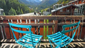 Ein Balkon mit zwei blauen Stühlen und Blick auf eine Berglandschaft mit Gebäuden und einem schneebedeckten Gipfel.
