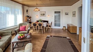 Una sala de estar acogedora con un sofá, una mesa de comedor con sillas, una mesa de centro con frutas y vasos, una chimenea y un amplio ventanal con cortinas.