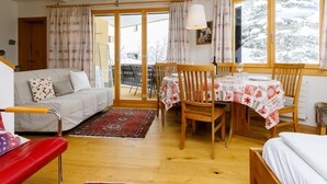 Ein gemütliches Zimmer mit einer Couch, Esstisch, Stühlen und Blick auf eine verschneite Landschaft durch große Fenster.