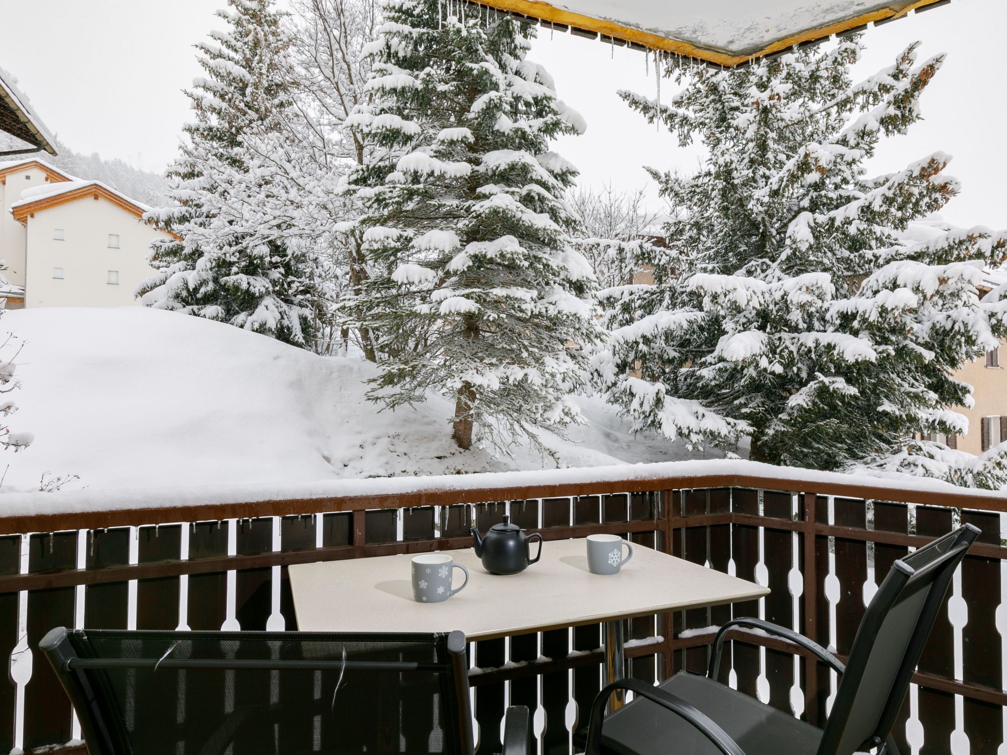 A snow-covered balcony with a table, chairs, and a teapot.
