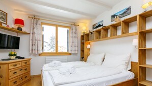 Ein Schlafzimmer mit einem hölzernen Bett, einem Fernseher, einem Schreibtisch mit Schubladen, einem Fenster mit Vorhängen und Regalen mit Dekorationselementen.