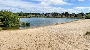 Una playa arenosa con huellas, un lago tranquilo y casas al fondo.