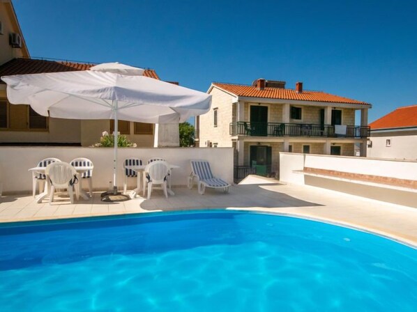 A swimming pool with white lounge chairs and an umbrella, surrounded by a building with a red-tiled roof and a balcony. - Apartment Orebić by Interhome (Orebić)