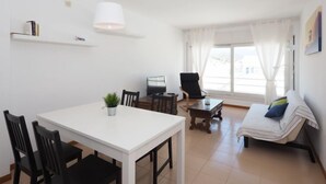 Un salón moderno con una mesa de comedor blanca, sillas negras, un televisor, un sillón negro, un sofá a rayas y un gran ventanal con cortinas.