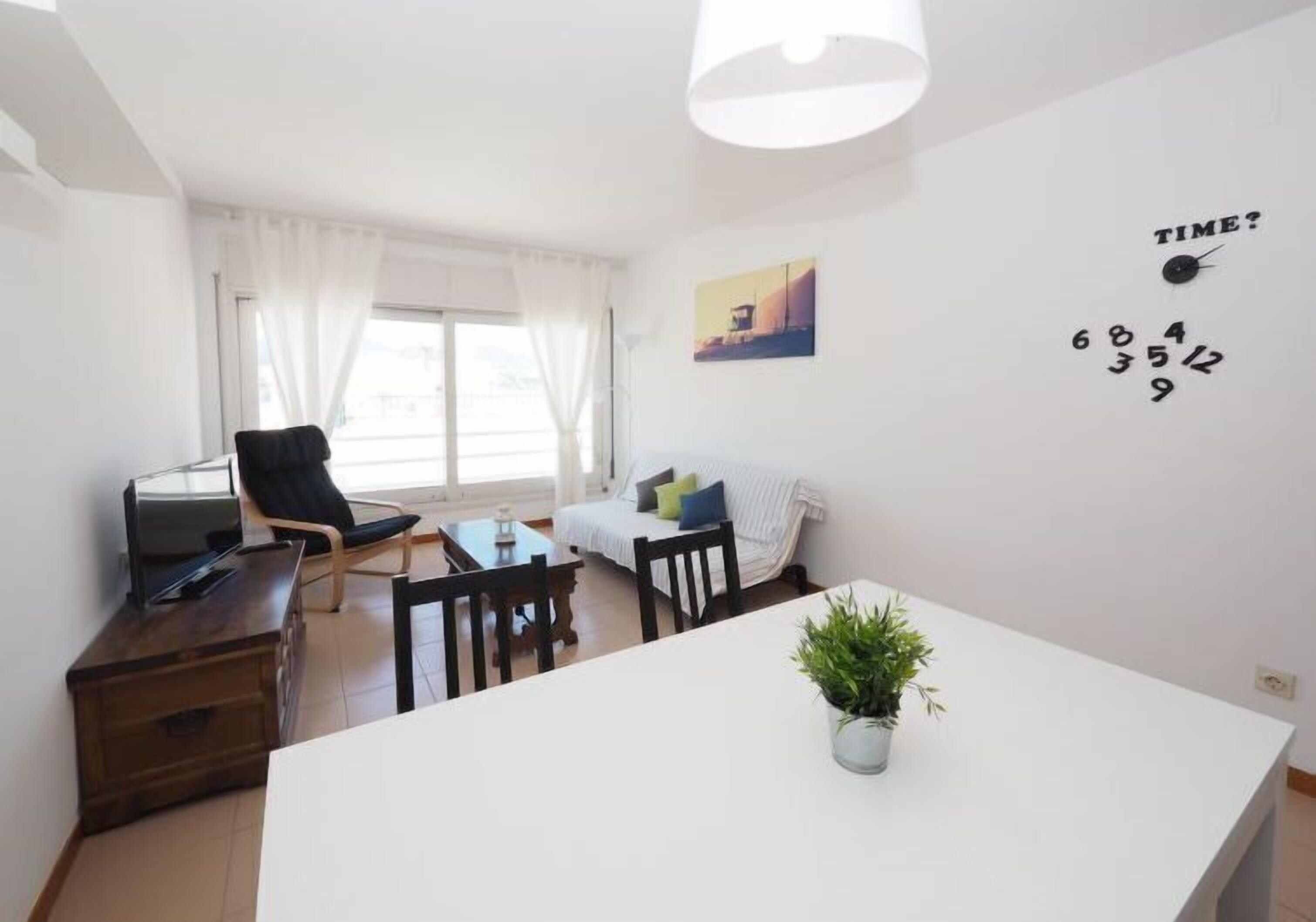 Un salon moderne avec un canapé blanc, une table à manger avec des chaises, un téléviseur posé sur un meuble en bois et une grande fenêtre avec des rideaux.