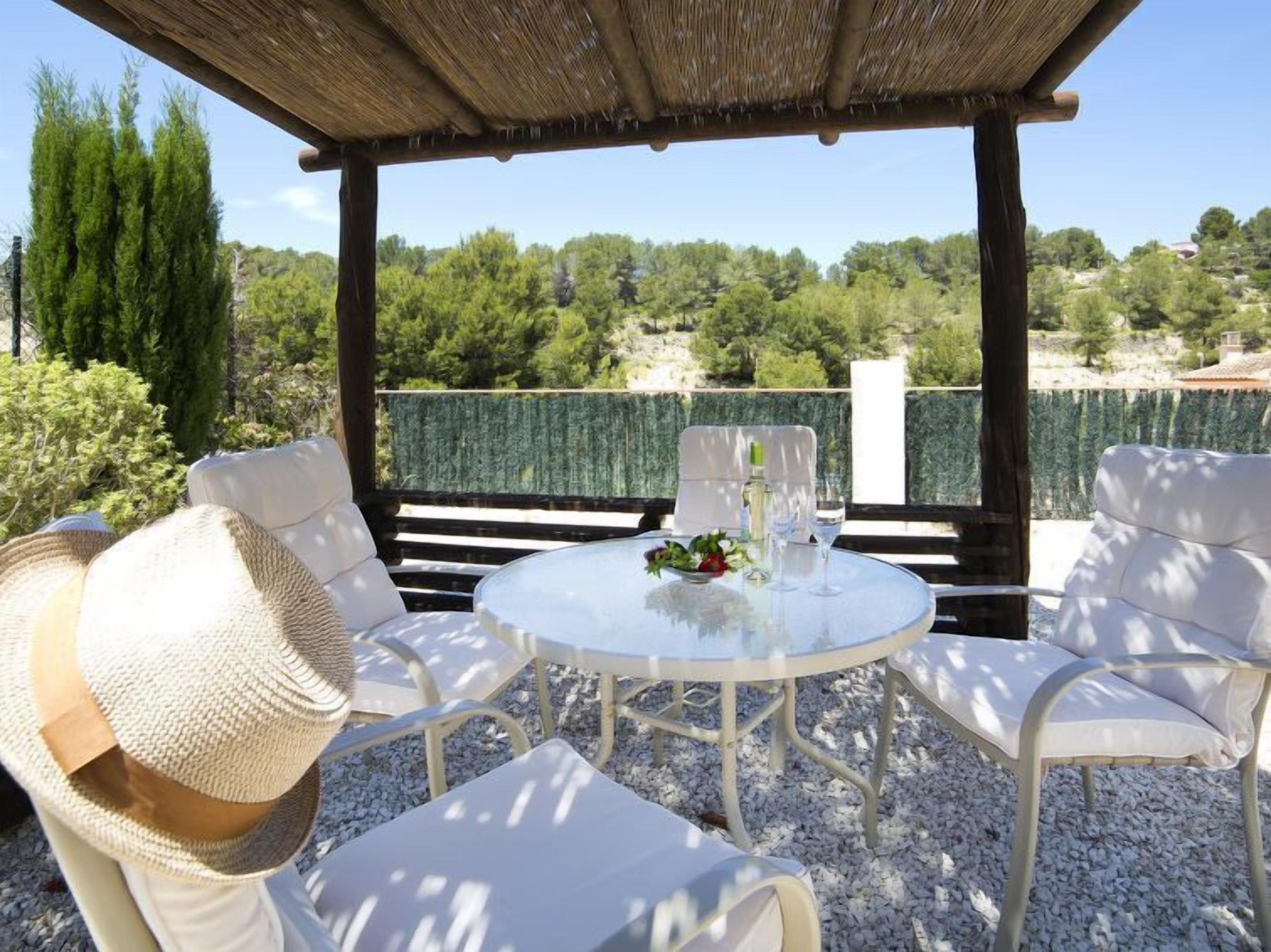 An outdoor seating area with white chairs and a round table, surrounded by a wooden pergola and a view of trees and hills.