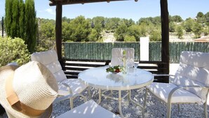 Ein Sitzbereich im Freien mit weißen Stühlen und einem runden Tisch, umgeben von einer hölzernen Pergola und Blick auf Bäume und Hügel.