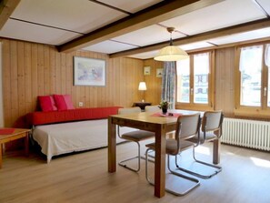 Quarto aconchegante com piso de madeira, uma cama com roupa de cama vermelha, uma mesa de jantar com cadeiras e uma janela com cortinas.