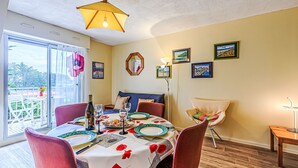 Ein Essbereich mit einem Tisch für zwei Personen, einer Couch und einem Sessel. Es gibt einen Balkon mit Blick auf Bäume und ein Gebäude.