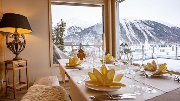 Une table à manger dressée pour six personnes, avec une vue sur les montagnes, une lampe et un plaid en fourrure.
