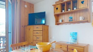 A wooden dining table with chairs, a television on a cabinet, and a shelf with decorative items.