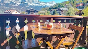 A wooden table and chairs on a balcony with a view of snow-capped mountains and houses in the background.