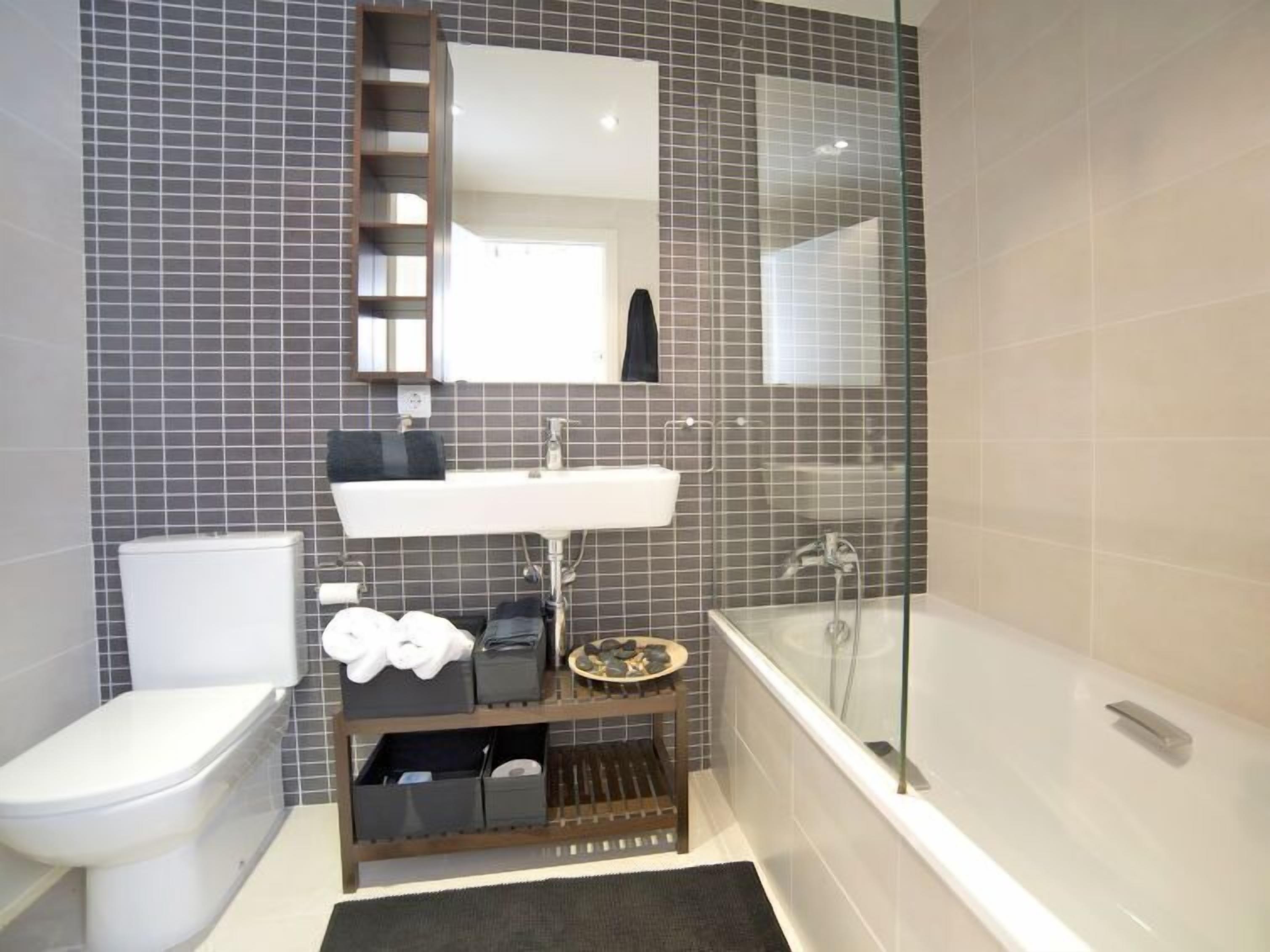 A modern bathroom with a toilet, sink, and shower. There is a mirror above the sink and a shelf with towels and toiletries.