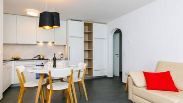 Une cuisine moderne avec des meubles blancs, une table à manger avec des chaises et un canapé agrémenté d’un coussin rouge.