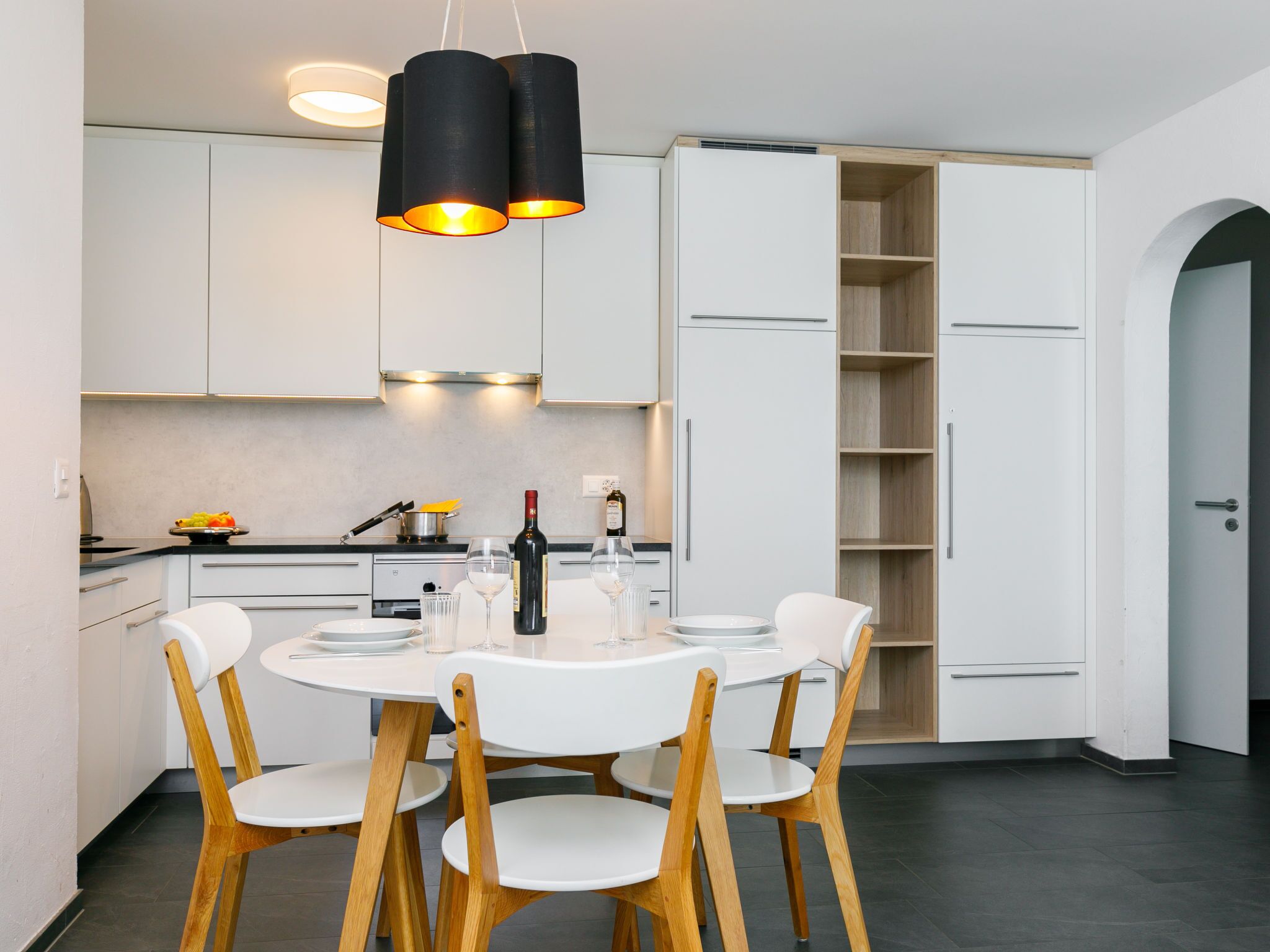 Une cuisine moderne avec des meubles blancs, une table à manger avec quatre chaises et une bouteille de vin posée sur le comptoir.