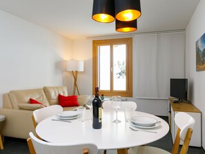 A dining area with a round table set for two, a sofa with red cushions, a lamp, and a TV on a wooden cabinet. - Residenza Chesa Margun 33-2 by Interhome (Silvaplana-Surlej)
