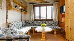 Ein gemütliches Blockhaus-Interieur mit Holzbalkendecke, einer Couch, einem runden Tisch und einem Fenster mit Vorhängen.