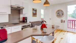 A modern kitchen with white cabinets, a built-in oven, a coffee machine, and a red kettle on the counter. There are two pendant lights hanging above the island, and a round mirror on the wall. - la Pergola by Interhome (Hossegor)