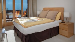Habitación de hotel con una cama grande, una mesita de noche con lámpara, una silla y vista al mar por la ventana.