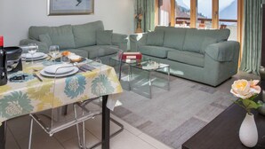 Una sala de estar con comedor, sofás verdes, una mesa de centro de vidrio y vistas a montañas a través de amplios ventanales.
