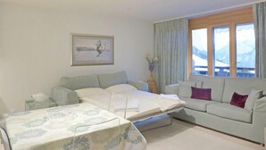 Una sala de estar con un sofá, una cama, una mesa con mantel, un cuadro enmarcado en la pared y una ventana con cortinas.