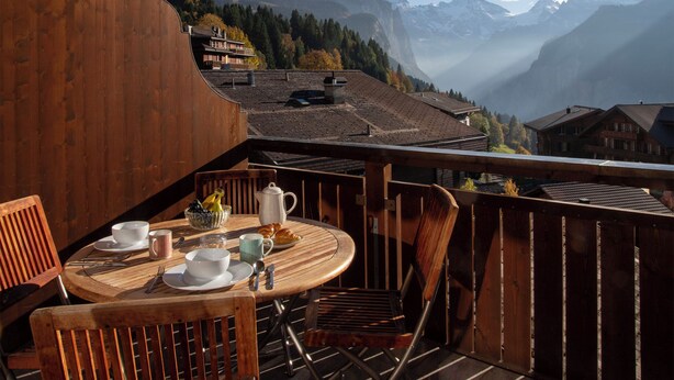 Una terraza de madera con una mesa lista para el desayuno, con vistas a un paisaje montañoso con casas y árboles.