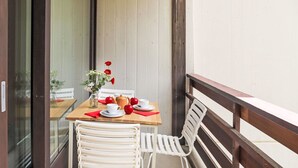 Un balcone con un tavolo di legno apparecchiato per due persone, sedie bianche, un vaso con fiori e un tovagliolo rosso.