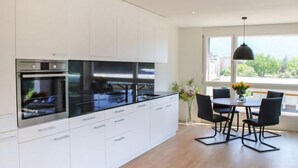 A modern kitchen with white cabinets, black countertops, and stainless steel appliances. There is a dining area with a table and chairs, and a large window providing natural light. - LaVille B-2-3 by Interhome (Locarno)