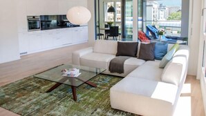 A modern living room with a glass coffee table, a sofa, and a kitchen area with white cabinets and appliances. - LaVille B-2-3 by Interhome (Locarno)