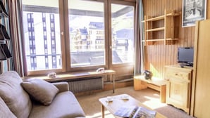 A modern living room with a sofa, a coffee table with a magazine, a TV on a wooden cabinet, and large windows offering a view of buildings. - Le Prariond (Val Claret) by Interhome (Tignes)