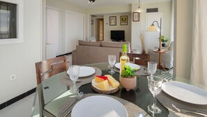 A dining area with a glass table set for two, a sofa in the background, and a television in the living room. - Les Roquetes by Interhome (Calpe/Calp)