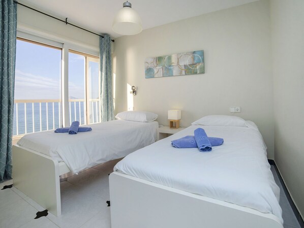 A hotel room with two beds, a large window with curtains, a wall-mounted lamp, and a painting.