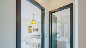 A modern bedroom with a bed, bedside table, and a hanging lamp. A bathroom with a shower and a mirror is visible through an open door. - Lucerna Due by Interhome (Ameglia)