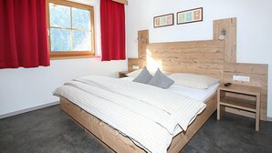 Een slaapkamer met een houten bedframe, een nachtkastje, een raam met rode gordijnen, en twee afbeeldingen in lijsten aan de muur.