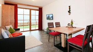 Een moderne woonkamer met een houten eettafel, rode fauteuils, een bank met kussens, een groot raam met rode gordijnen en een wandtelevisie.
