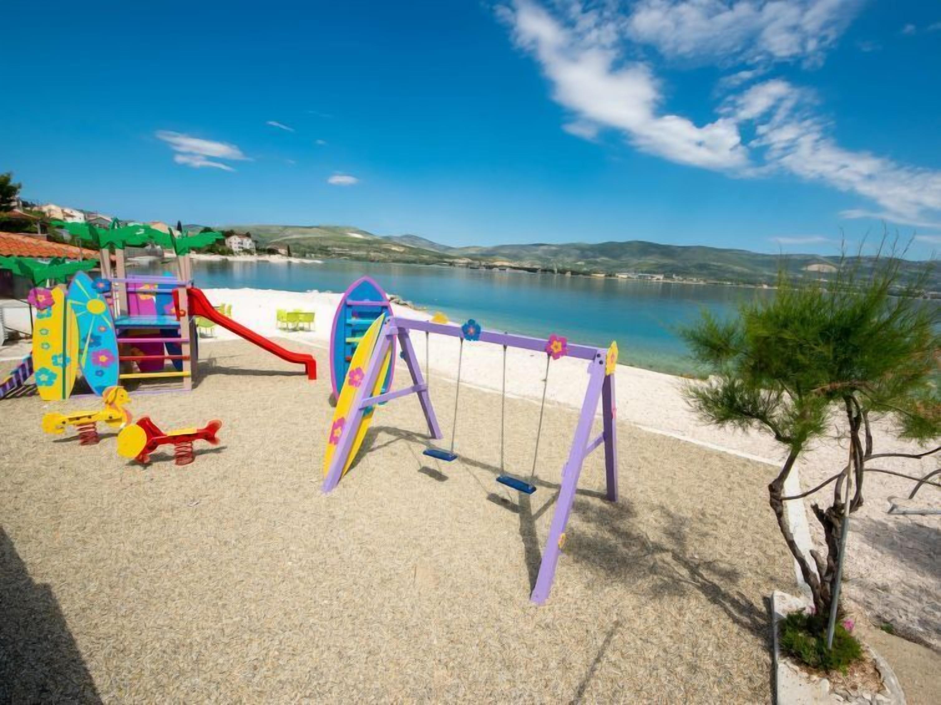 A playground with swings and slides near a beach with clear blue water and hills in the background.