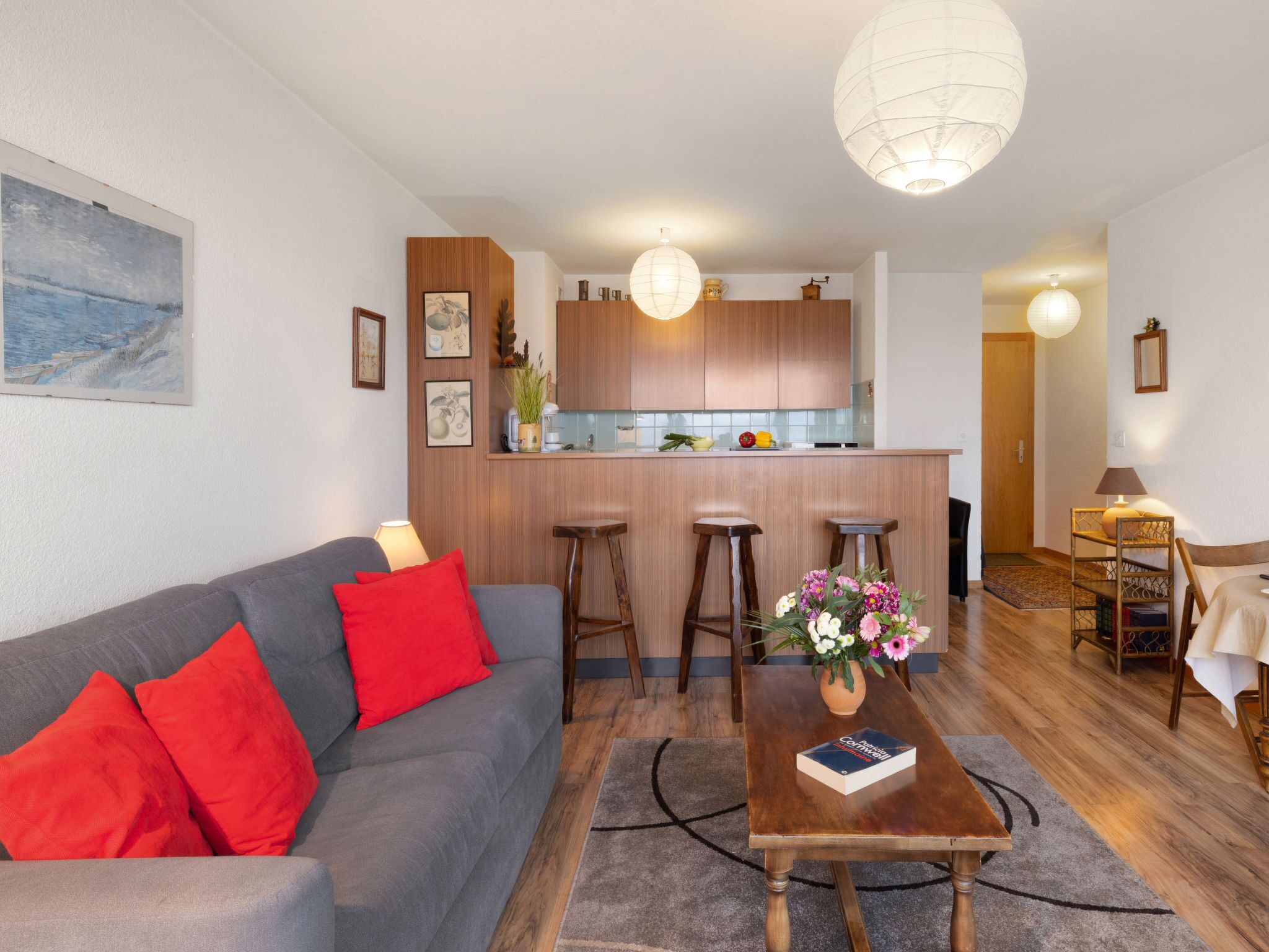A modern living room with a grey sofa, red cushions, a coffee table with a book, wooden flooring, and a kitchen area with wooden cabinets and bar stools.