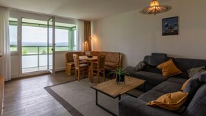 A modern living room with a sofa, a dining table with chairs, a coffee table, and a large window offering a view of the outdoors. - Apartment K 40 by Interhome (Dittishausen)