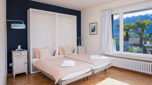 A bedroom with two beds, a nightstand, a lamp, a window with a view of mountains, and a radiator. - Suite Classic by Interhome (Ascona)