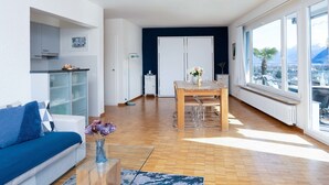A modern living room with a sofa, a dining table with chairs, a kitchen area, and large windows offering a view of the city and mountains. - Suite Classic by Interhome (Ascona)